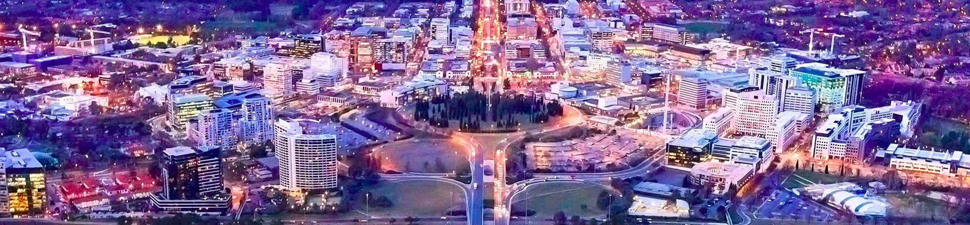 Canberra from the air at dusk: the city centre lit up, with roads running out from a ring of trees.